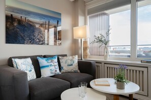 Een moderne woonkamer met een grijze bank, drie decoratieve kussens, een ronde witte tafel met een boek en een potplant, een vloerlamp en een groot schilderij van een strandgezicht.