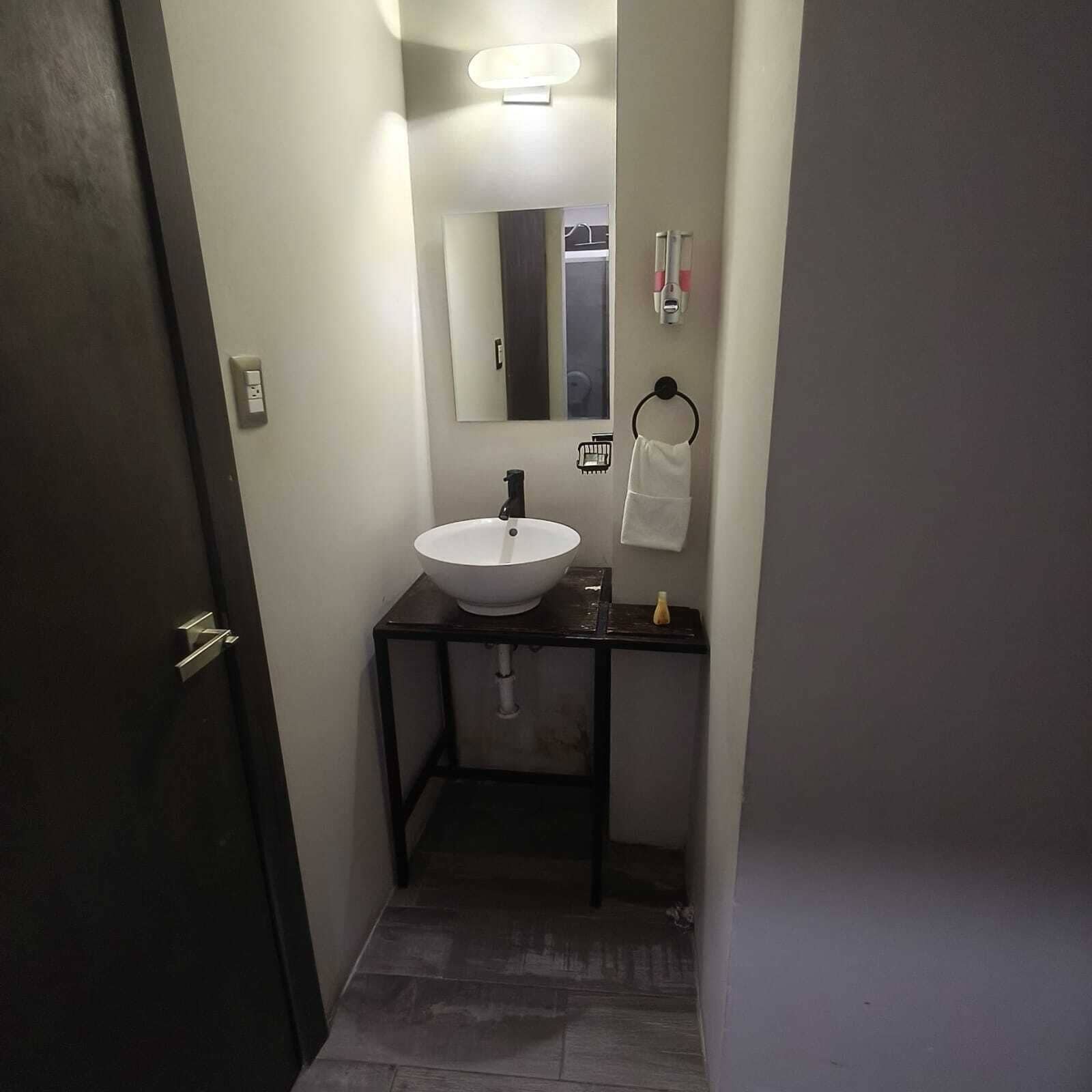 Un couloir étroit mène à une salle de bain équipée d’un lavabo blanc, d’un miroir et d’un porte-serviettes.