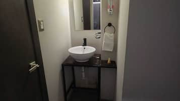 Un couloir étroit mène à une salle de bain équipée d’un lavabo blanc, d’un miroir et d’un porte-serviettes.