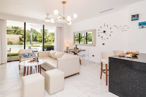 Un salon moderne avec un canapé, une table basse et un coin repas. Une grande fenêtre offre une vue sur une piscine et sur la verdure environnante.