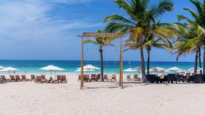 A beach with lounge chairs and umbrellas, a sign for the Cana Bay Beach Club, and palm trees. - Directo piscina jardín terraza @ Cana Rock (Bavaro)
