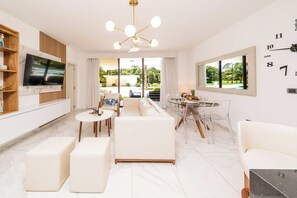 A modern living room with a flat-screen TV, white sofas, a dining area with a round table, and a large window with a view of a pool and greenery. - Aqua Joy | 3BR Family Swim-Up + Whirlpool (Bavaro)