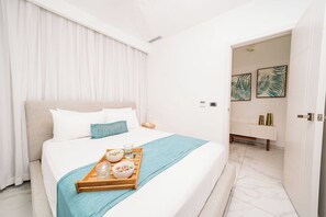A hotel room with a bed, a tray with breakfast items, a white bathroom with a vanity, and artwork on the wall. - Aqua Joy | 3BR Family Swim-Up + Whirlpool (Bavaro)