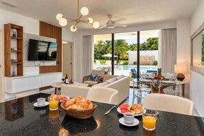 A modern hotel room with a dining area, a sofa, a TV, and a view of a swimming pool.