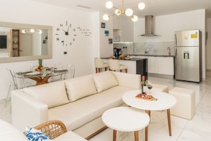 Un salon moderne comprenant un canapé, deux tables rondes, un coin cuisine avec un réfrigérateur et une horloge murale.