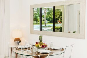 A dining table with clear glass top, surrounded by chairs, set with plates, glasses, and a fruit bowl. A lamp is on the table, and a large mirror reflects the outdoor view. - Aqua Joy | 3BR Family Swim-Up + Whirlpool (Bavaro)