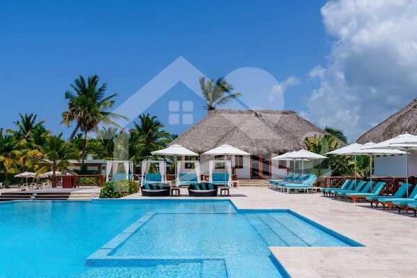 A pool area with lounge chairs and umbrellas, surrounded by palm trees and thatched-roof structures.