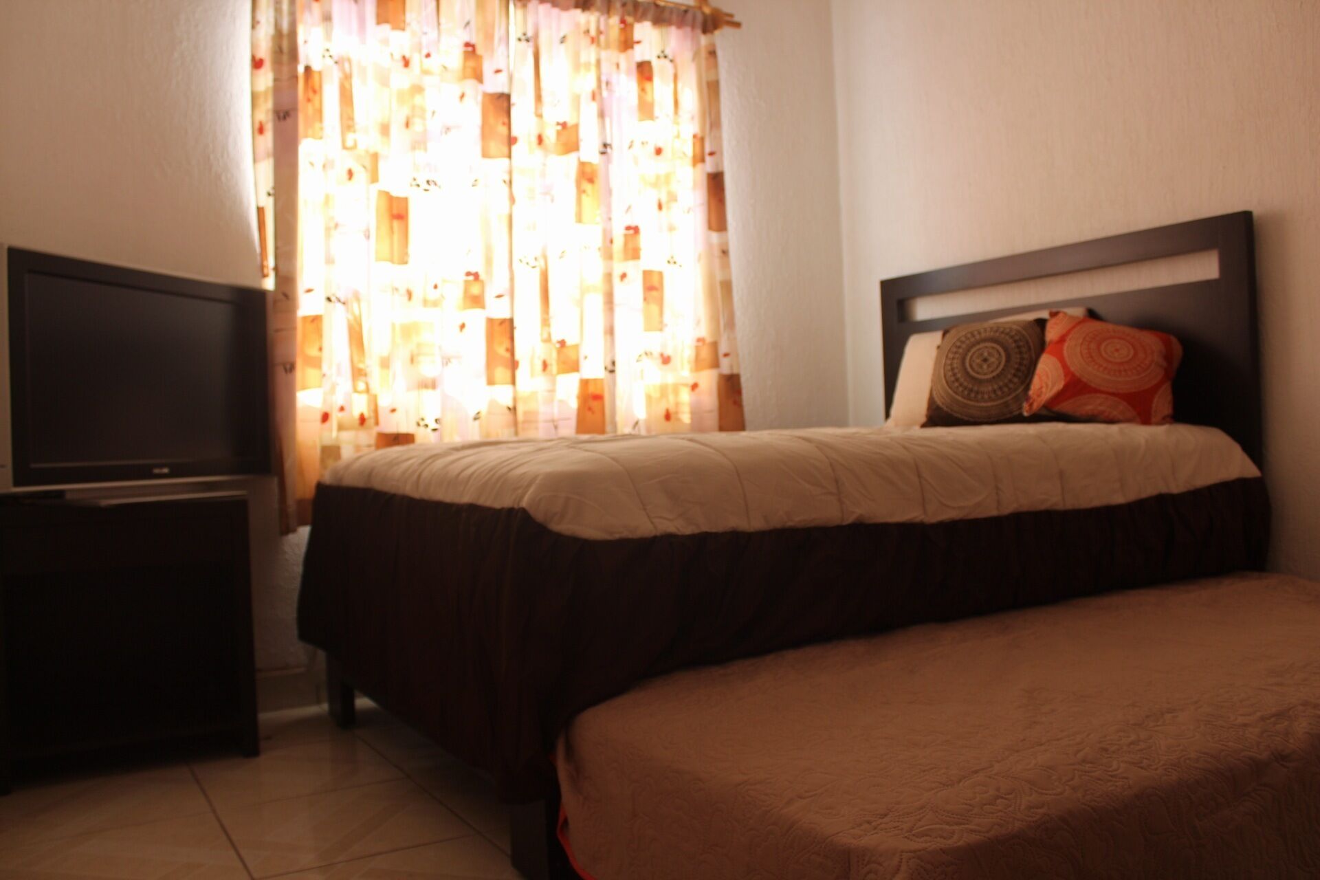 A hotel room with a bed, a TV, and a window with patterned curtains.