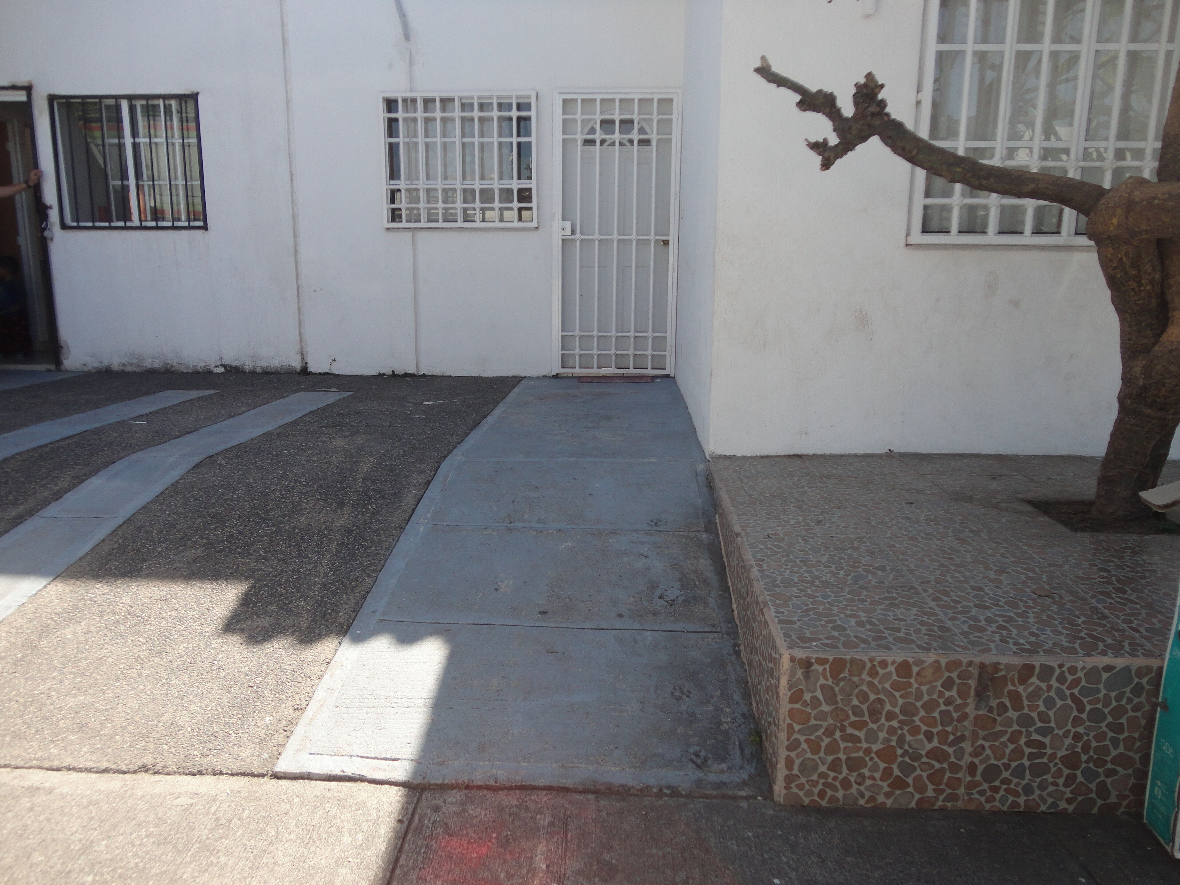 Esterno di un edificio con ingresso pavimentato, porta con cancello e area piastrellata con motivo in pietra.