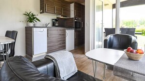 A modern kitchen with wooden cabinets, a built-in oven, and a coffee machine. There is a dining area with a table and chairs, and a sofa with a blanket. A bowl of fruit is on the table. - 6 Person Holiday Park Home in Asperup (Middelfart)