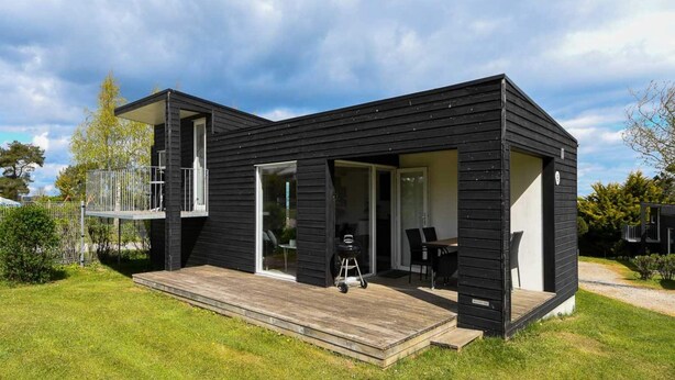 A modern black house with a wooden deck, a sliding glass door, and a patio area with outdoor furniture. - 6 Person Holiday Park Home in Asperup (Middelfart)