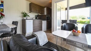 A modern kitchen with wooden cabinets, a white refrigerator, and a coffee maker. There is a dining area with a table and chairs, and a sofa with a bowl of fruit on it. - 6 Person Holiday Park Home in Asperup (Middelfart)