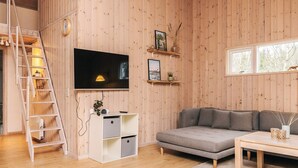 Una sala de estar acogedora con detalles de madera, un televisor en la pared, un sofá gris, una mesita pequeña y una escalera que sube a un nivel superior.