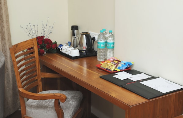 A wooden desk with a chair, a coffee maker, two water bottles, a tray of snacks, and a vase of red roses. - Sai Neem Tree Hotel (Rahata)