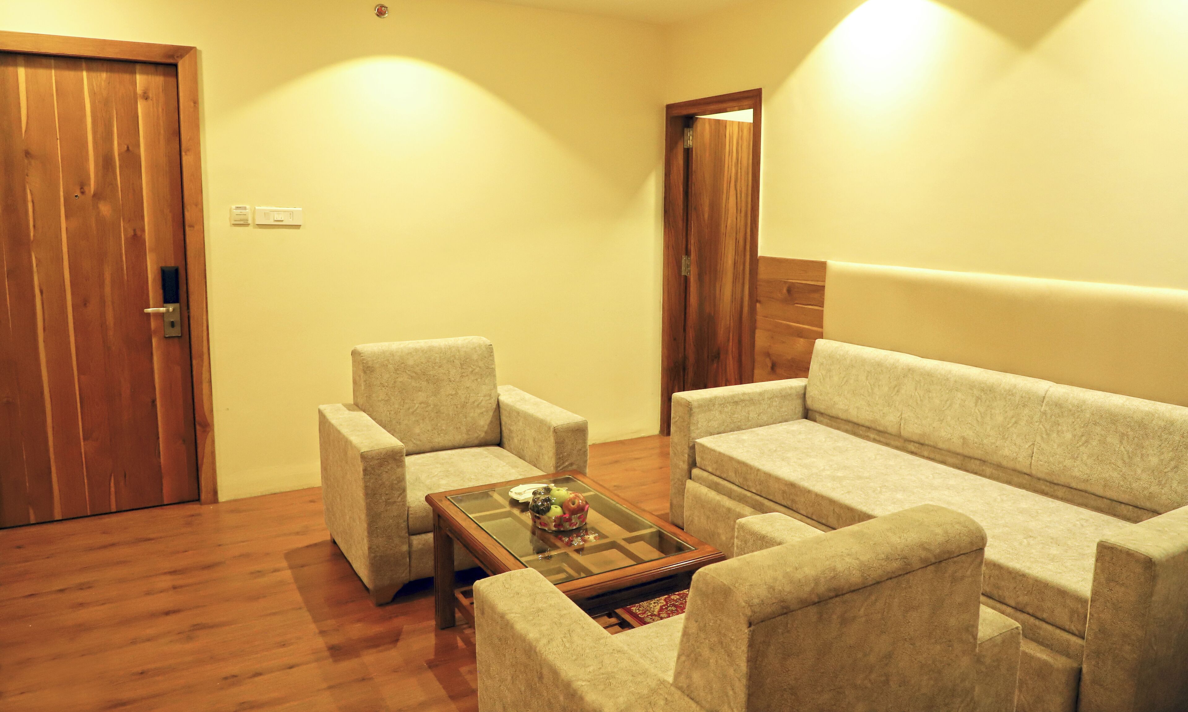 A room with a wooden door, a beige sofa, two armchairs, and a glass coffee table.