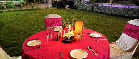 A round table set outdoors with a red tablecloth, white plates, and silverware.