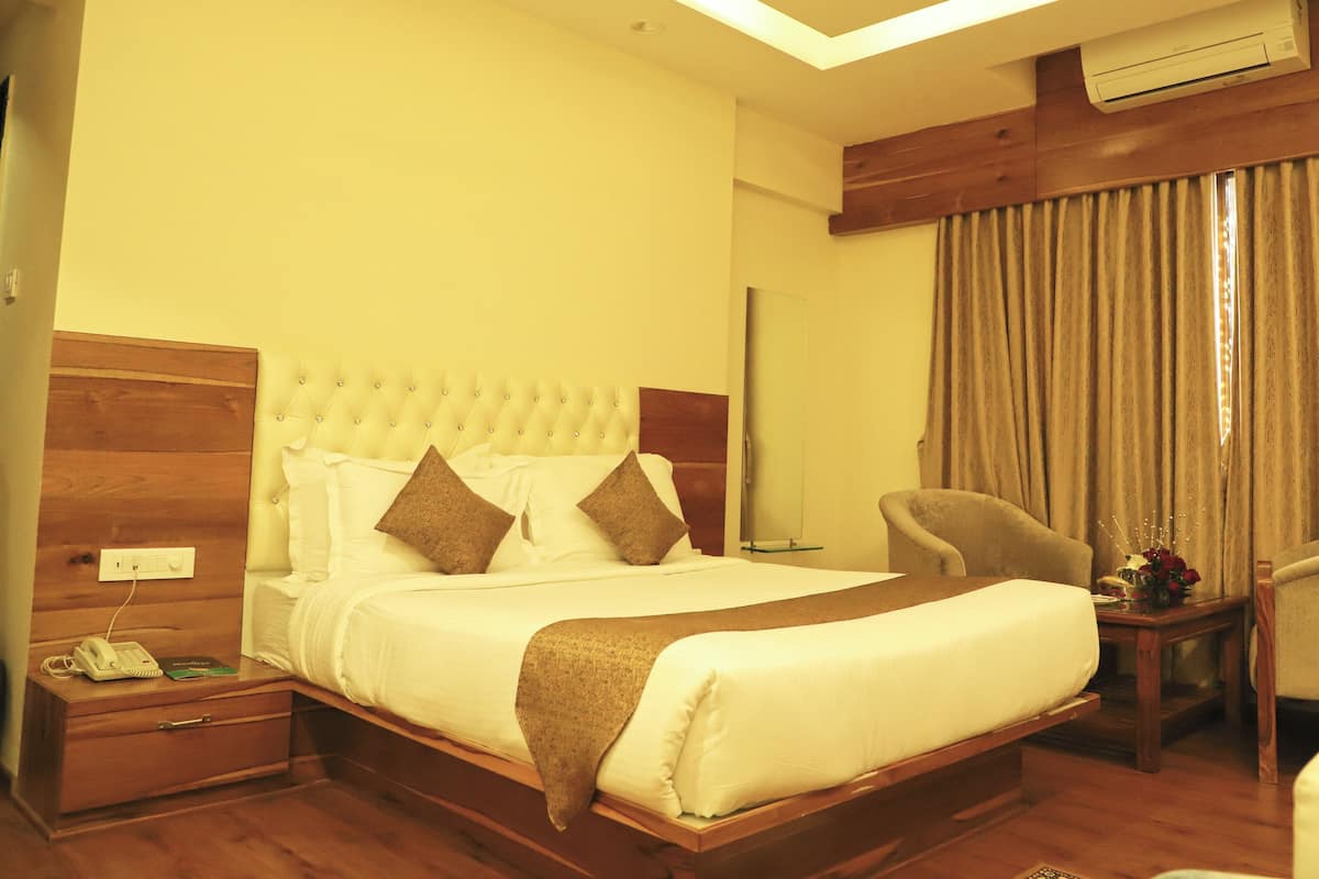 A hotel room with a large bed, a wooden headboard, a bedside table with a phone and a book, a chair, and a window with curtains.