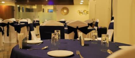 Un salón de banquetes con mesas redondas dispuestas para un evento formal, con manteles blancos, cubiertos dorados y cintas de color azul marino en las sillas.