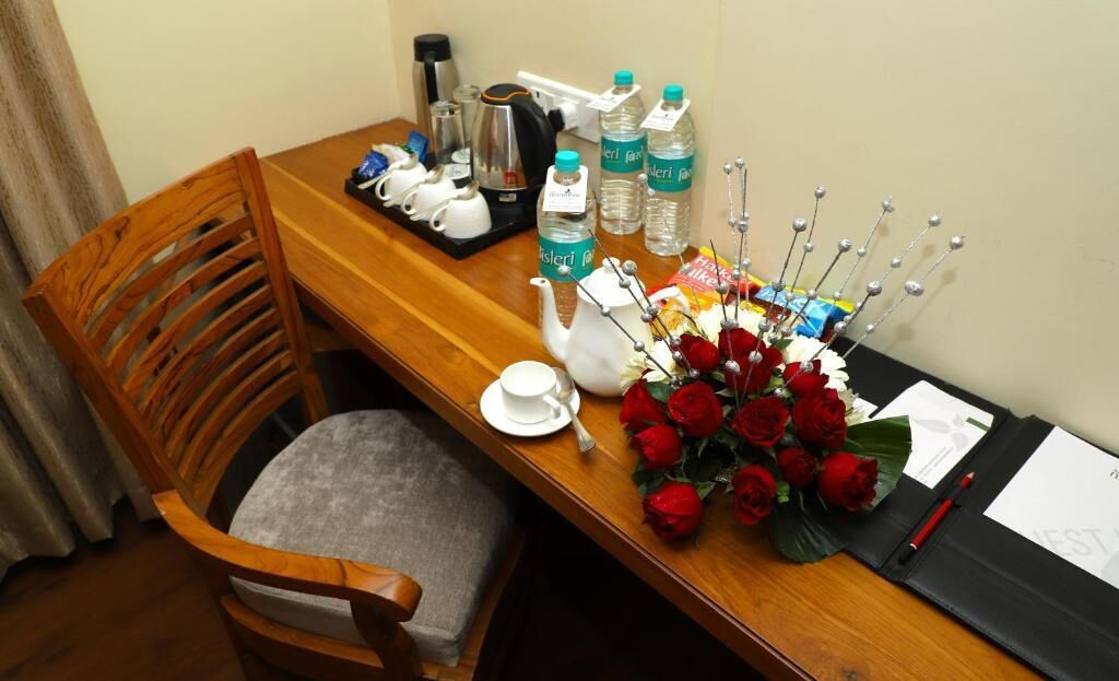 Une table de salle à manger en bois, sur laquelle se trouvent une théière blanche, des tasses, une bouilloire et des bouteilles d’eau. On peut également y voir des roses rouges et un dossier noir contenant des documents.