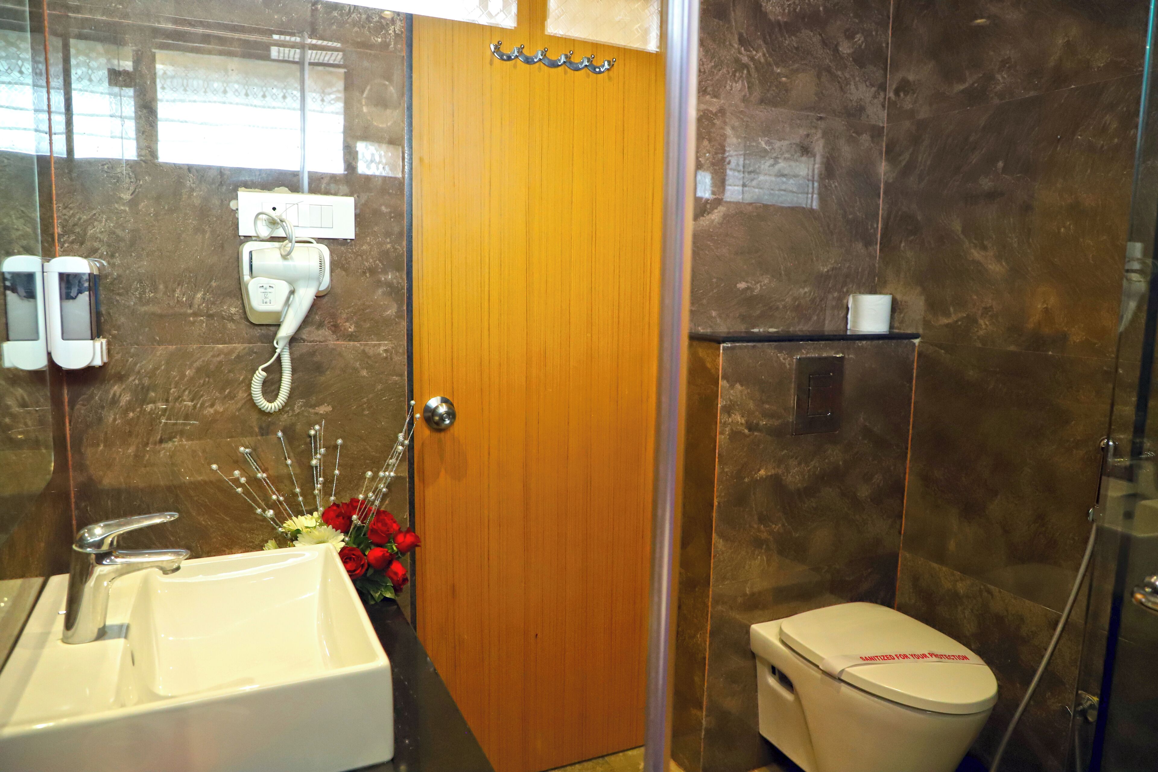 Un baño con puerta de madera, un lavamanos blanco con espejo, un secador de pelo y un inodoro con etiqueta roja.