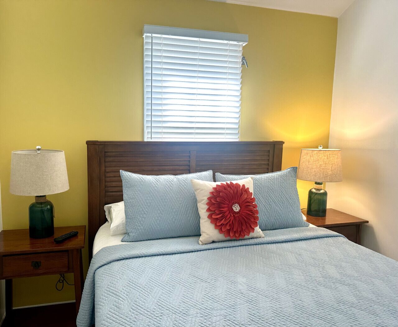 A neatly made bed with a blue quilt, two pillows, and a red decorative pillow. There are two bedside lamps on wooden nightstands, and a window with blinds in the background.
