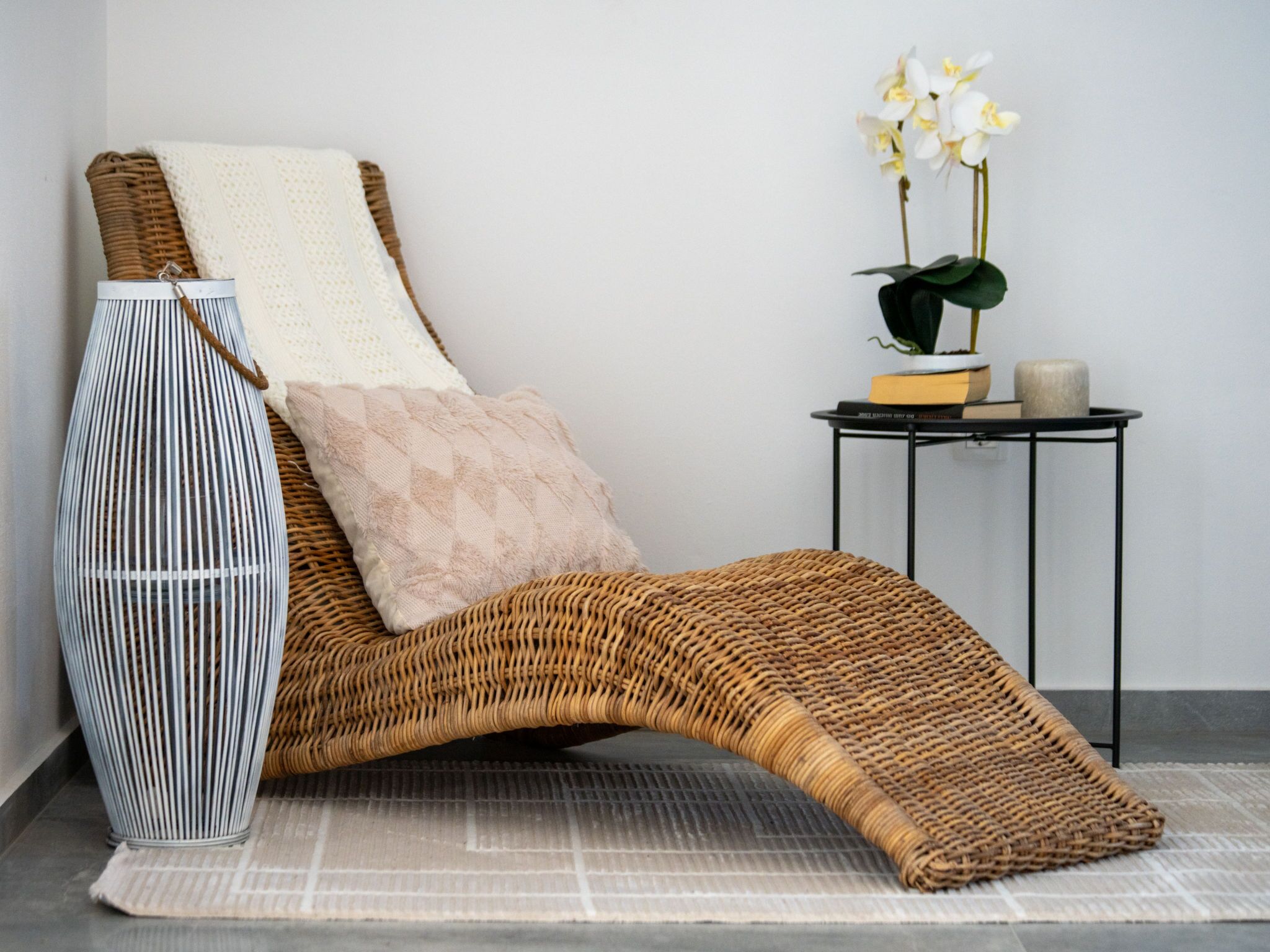 A wicker lounge chair with a cushion, a tall striped vase, and a small table with a potted plant and books.