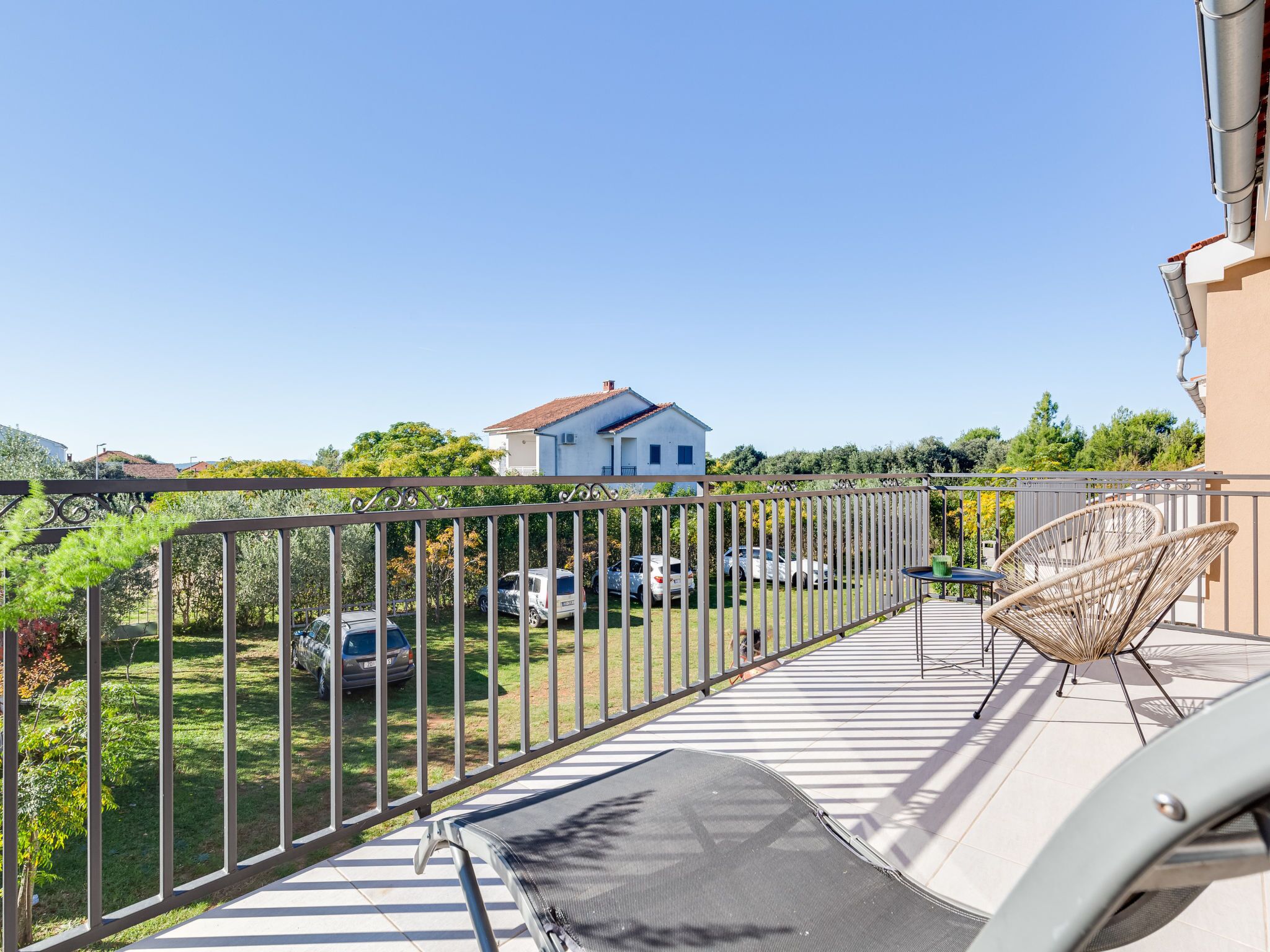 A balcony with a metal railing, a wicker chair, and a small table. In the background, there is a house and parked cars.