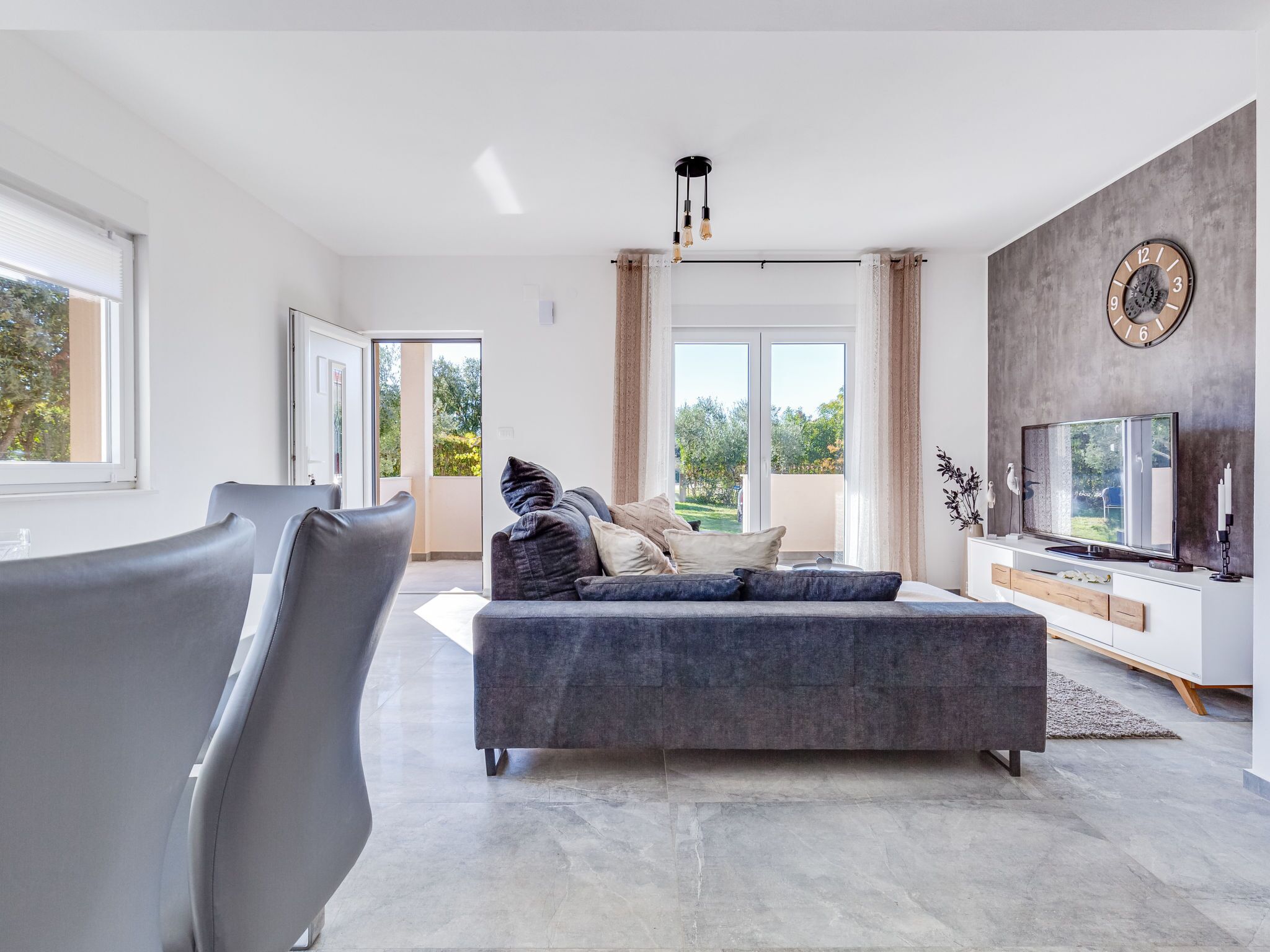 A modern living room with a grey sofa, a TV, and a large window offering a view of greenery.