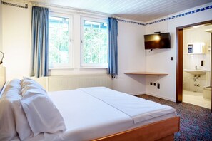 Ein Hotelzimmer mit Bett, Fernseher, Fenster mit Vorhängen, einer kleinen Ablagefläche und einem durchgegangene Tür sichtbaren Badezimmer.