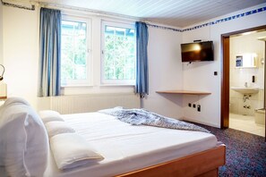 Habitación de hotel con cama, televisión, una pequeña estantería y baño visible a través de una puerta abierta.