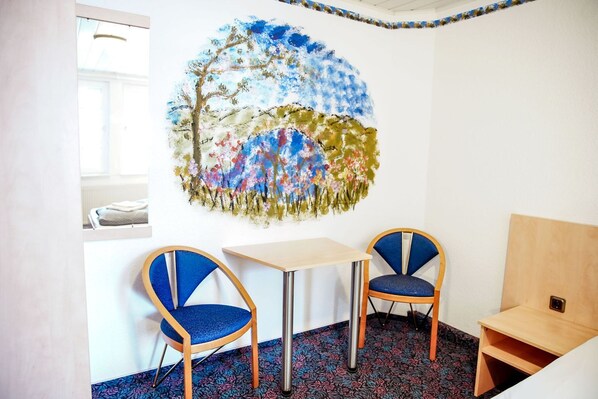 A room with a large abstract painting on the wall, two chairs with blue upholstery, a small table, and a wooden bench.