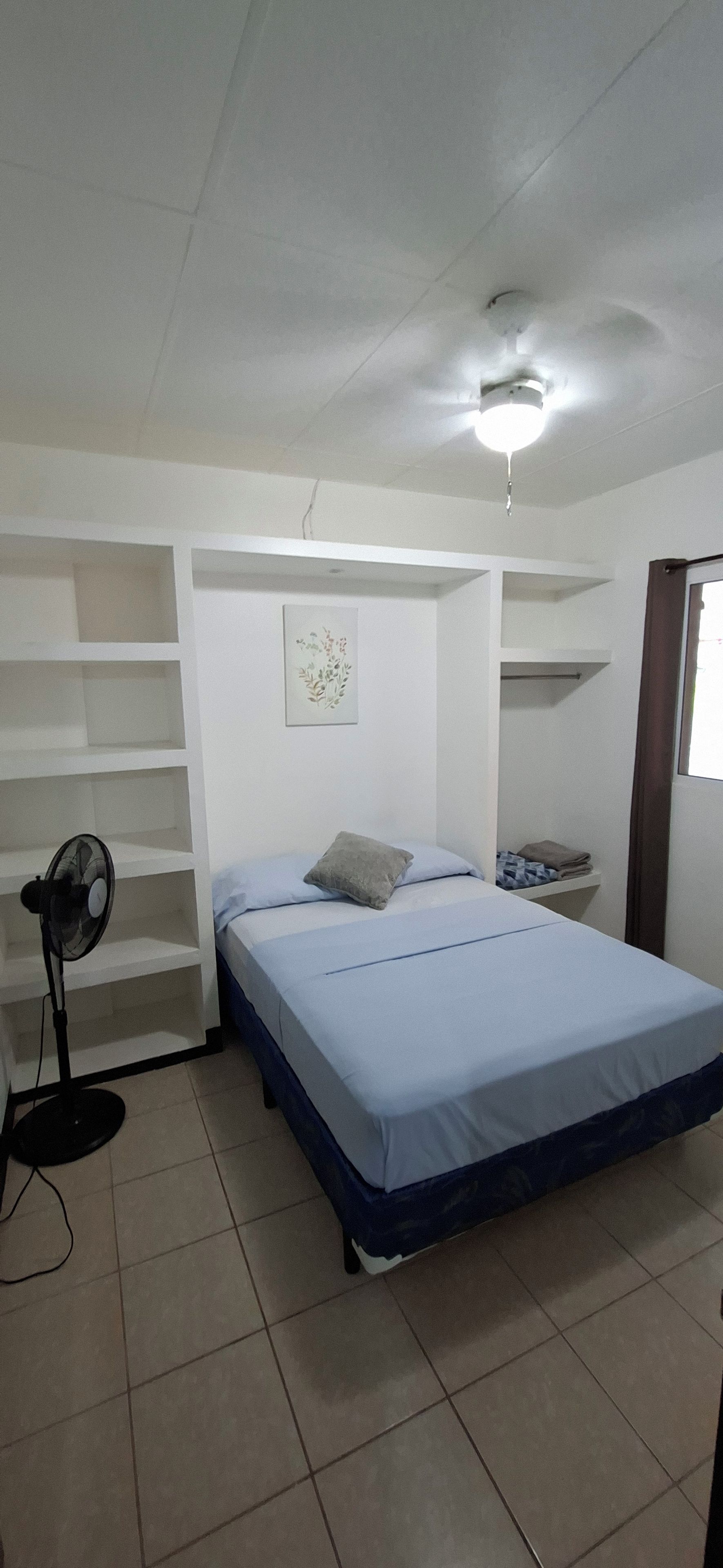 A single-bed room with a ceiling fan, a bedside table, a small shelf, and a window with curtains.