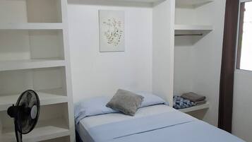 A single-bed room with a ceiling fan, a bedside table, a small shelf, and a window with curtains.