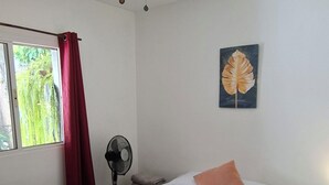 Una camera da letto con un letto, una ventola a soffitto, una finestra con tende, una ventola a parete e un quadro.