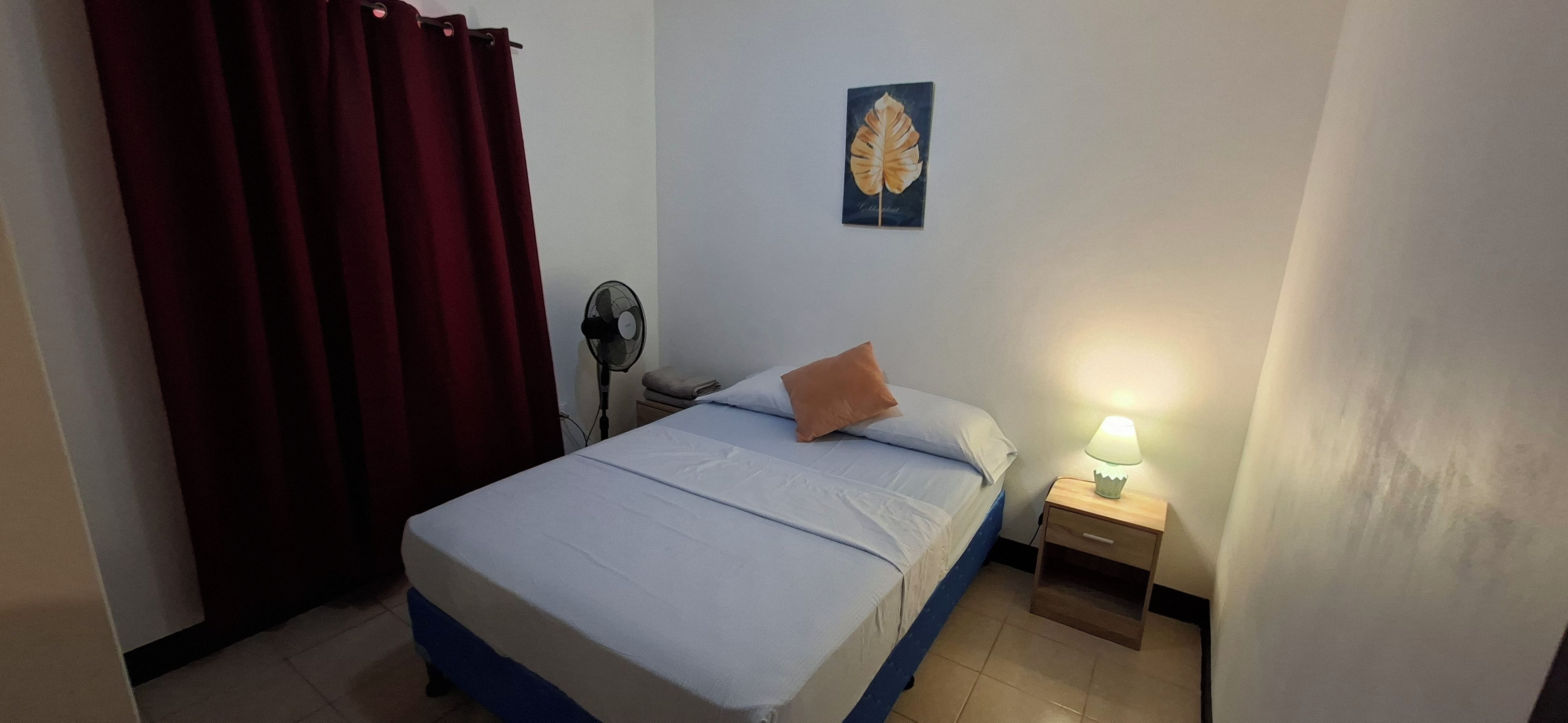 Un dormitorio con una cama, una mesita de noche, una lámpara, un ventilador y un cuadro en la pared.