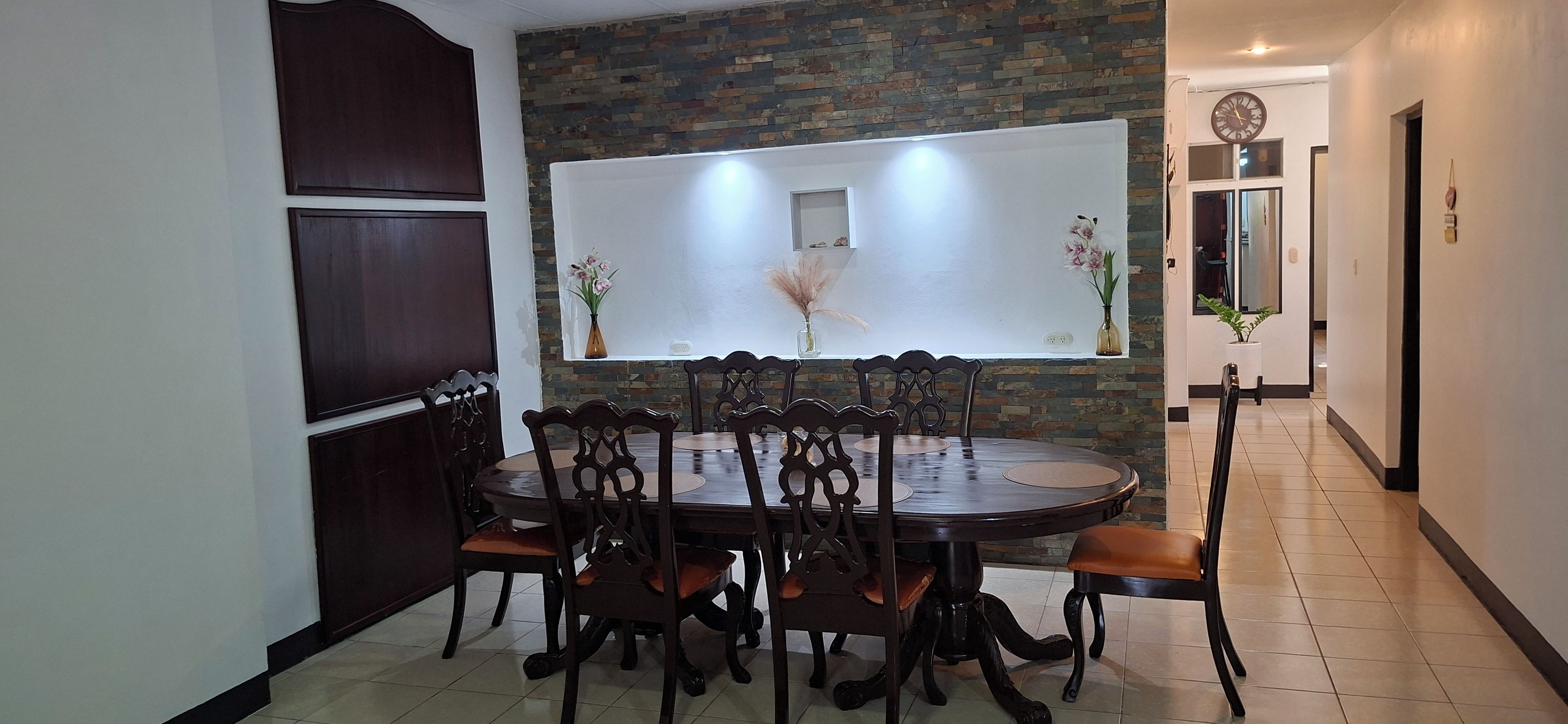 Un comedor con una mesa redonda, seis sillas y una pared decorativa con una ventana arriba.