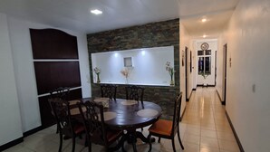 Zona pranzo con tavolo rotondo, sedie, TV a parete e corridoio che porta a una porta con orologio.