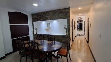 A dining area with a round table, chairs, a wall-mounted TV, and a hallway leading to a door with a clock.