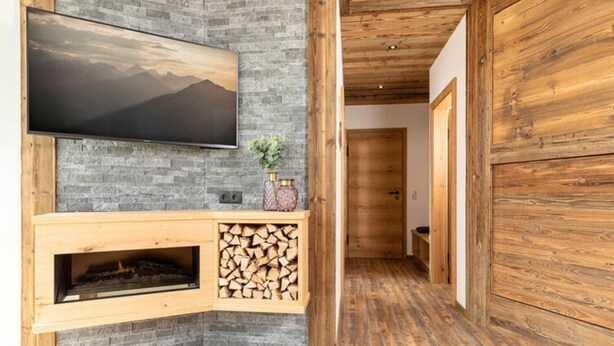 A modern living room with a stone fireplace, a mounted TV, and a wooden door. - Apartment in Fugen Near Spieljoch Ski Lift (Fuegen)
