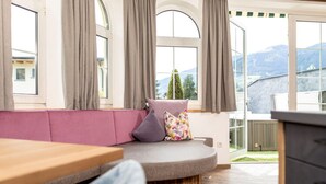A cozy seating area with a cushioned bench, a wooden table, and a view of a mountain landscape through large windows with curtains. - Apartment in Fugen Near Spieljoch Ski Lift (Fuegen)