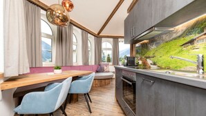 A modern kitchen with a wooden countertop, two blue chairs, a built-in oven, and a view of a green hillside through a large window. - Apartment in Fugen Near Spieljoch Ski Lift (Fuegen)