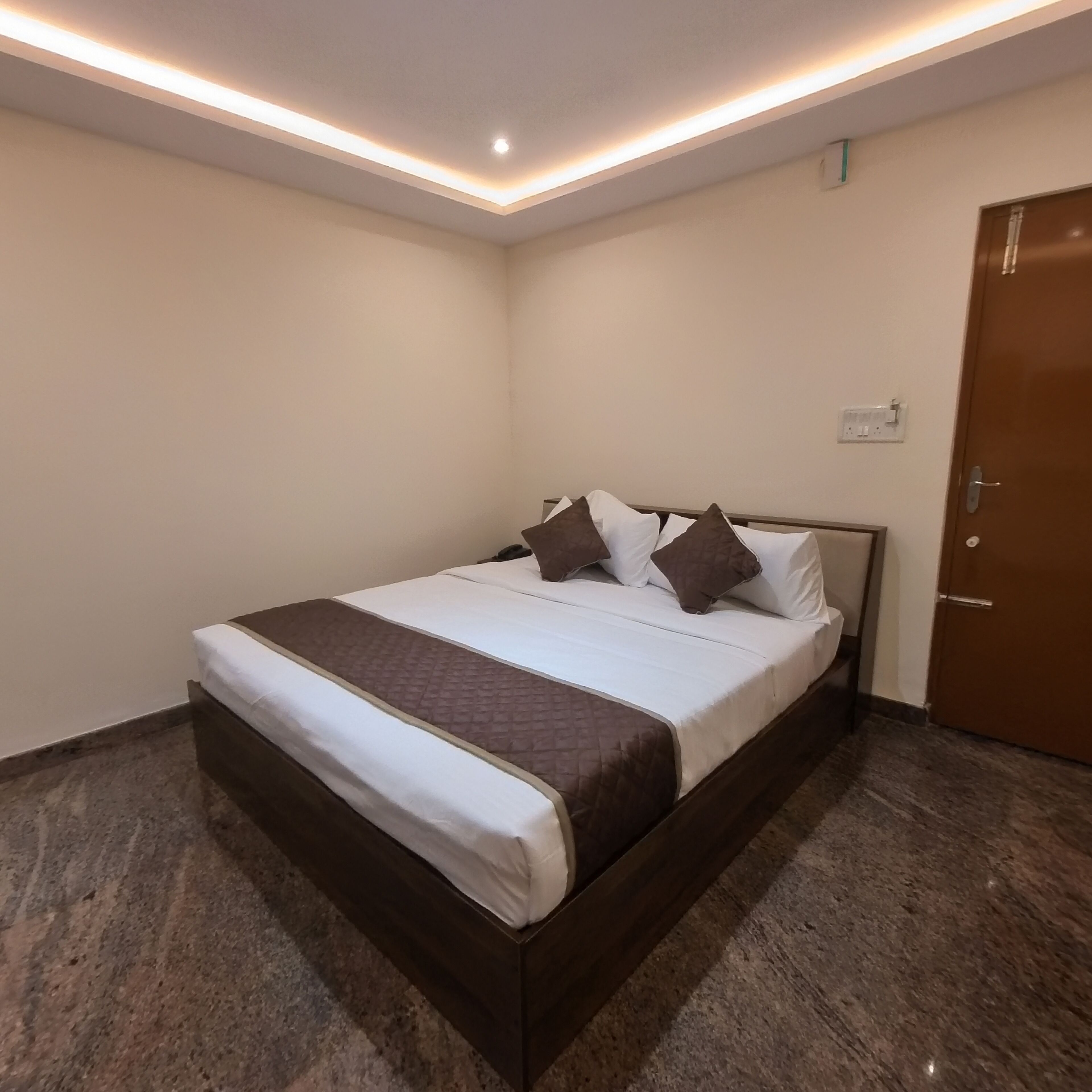 A neatly made bed with a brown headboard and white bedding, a wooden floor, and a door on the right.