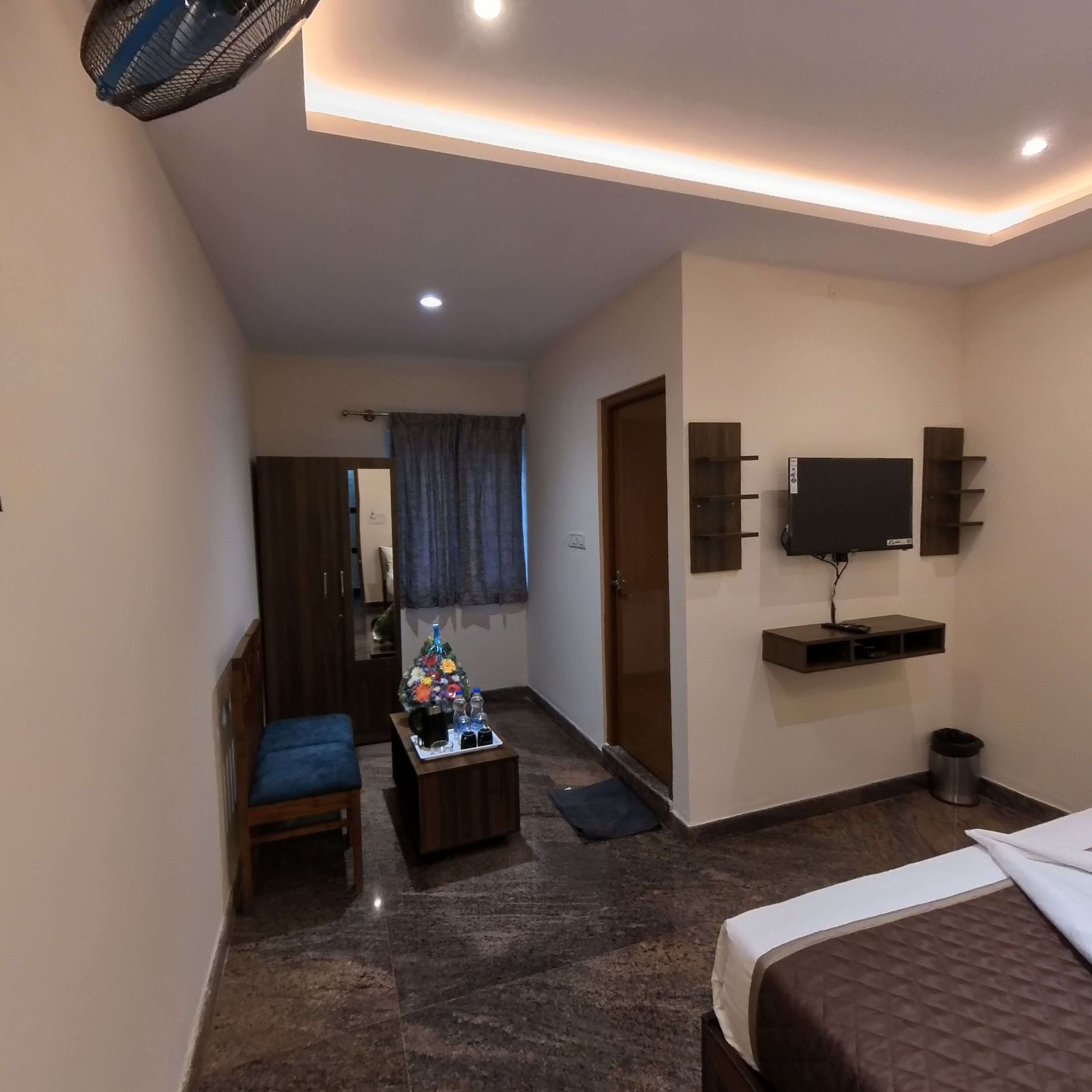 A hotel room with a bed, a small seating area with a chair and table, a TV mounted on the wall, and a door leading to another room.