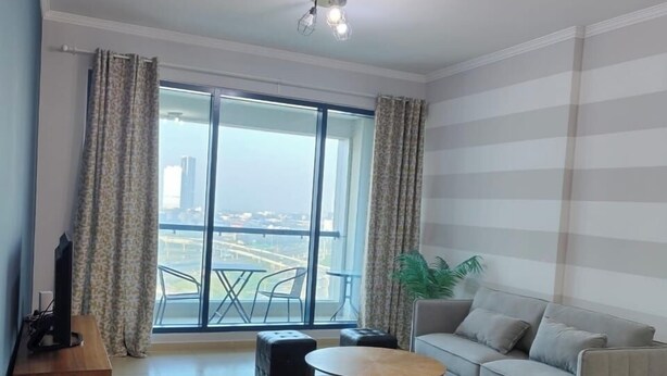 A modern living room with a large window, a grey sofa, a coffee table, and a TV on a wooden stand. - Charming and Comfortable 1BR | Jumeirah Bay X1| JLT (Dubai)