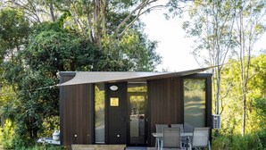 A modern cabin with a dark exterior, a glass door, and a gravel patio with two chairs.