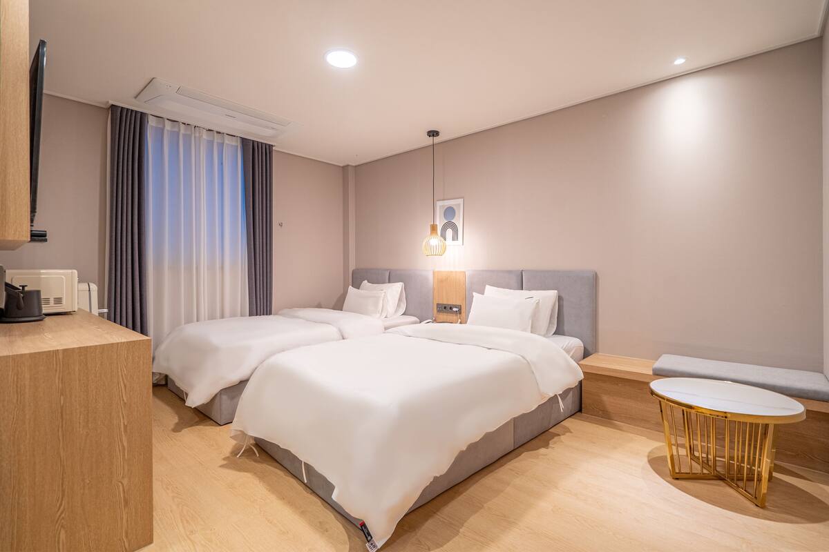 Una habitación de hotel moderna con dos camas, un escritorio de madera, una pequeña mesa redonda y un banco.