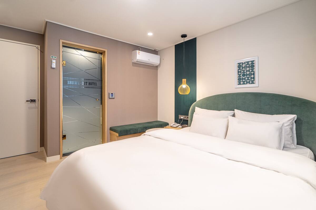 Habitación de hotel con cama, mesita de noche, cabecera verde, aire acondicionado de pared, cuadro enmarcado y puerta con panel de vidrio.