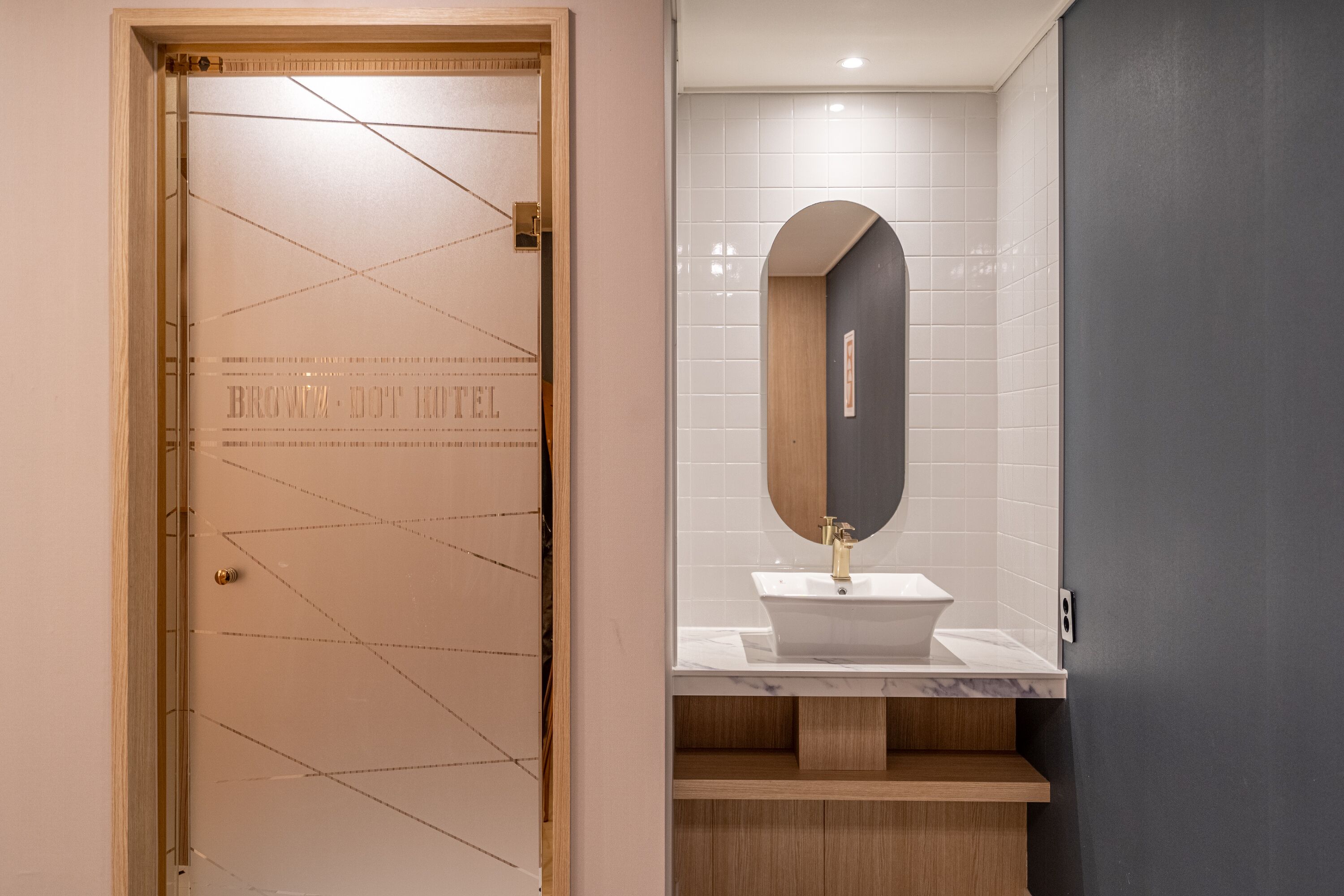 Una camera d'hotel con una porta in legno, una parete rivestita di piastrelle bianche, un piano di lavoro in marmo e un mobiletto a specchio in legno.