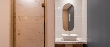 A hotel room with a wooden door, a white tiled wall, a marble countertop, and a wooden vanity.