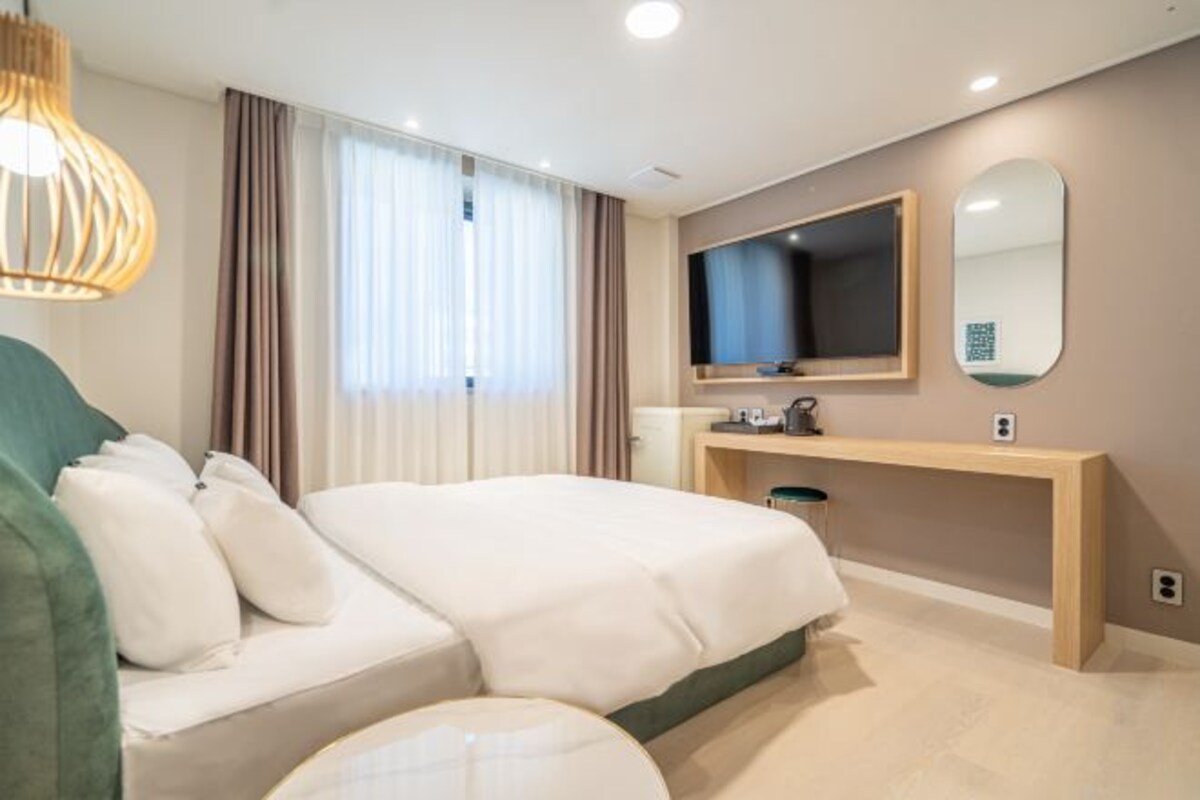 Una habitación de hotel moderna con una cama, mesita de noche, espejo y un amplio ventanal con cortinas.